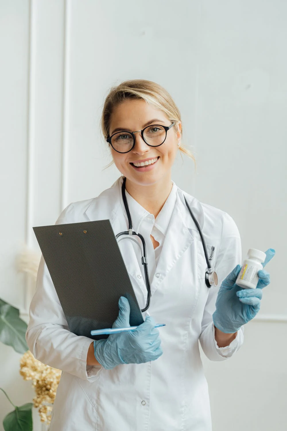 a young woman in a lab coat is holding a bottle of pills and a clipboard, she has a big smile as her expression.