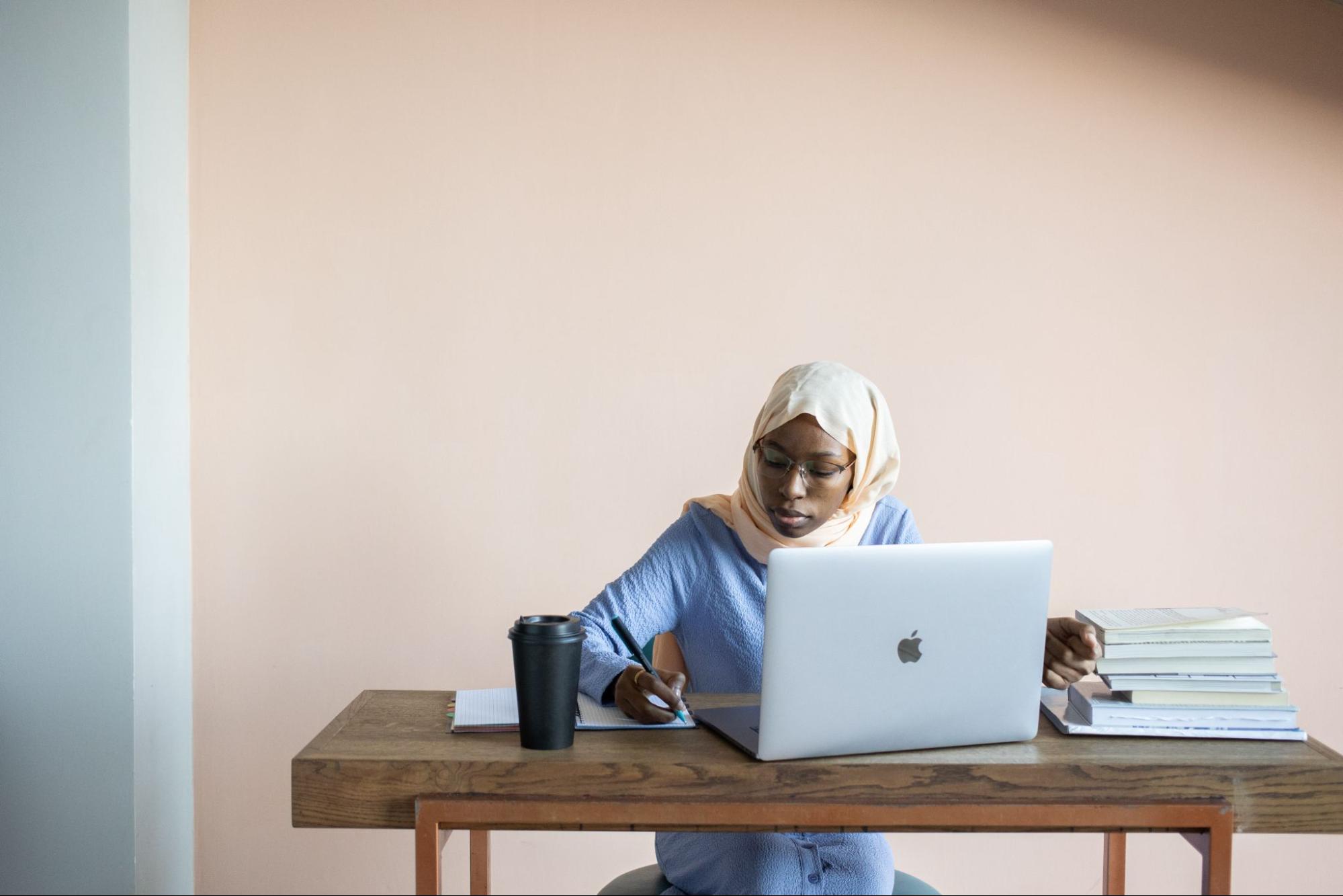 A woman sits in front of a laptop computer while she takes notes on a notepad