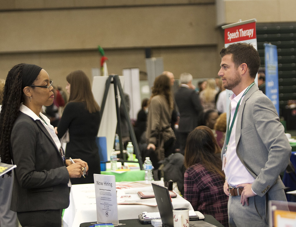 A man and a woman stand facing one another over a table with hiring materials