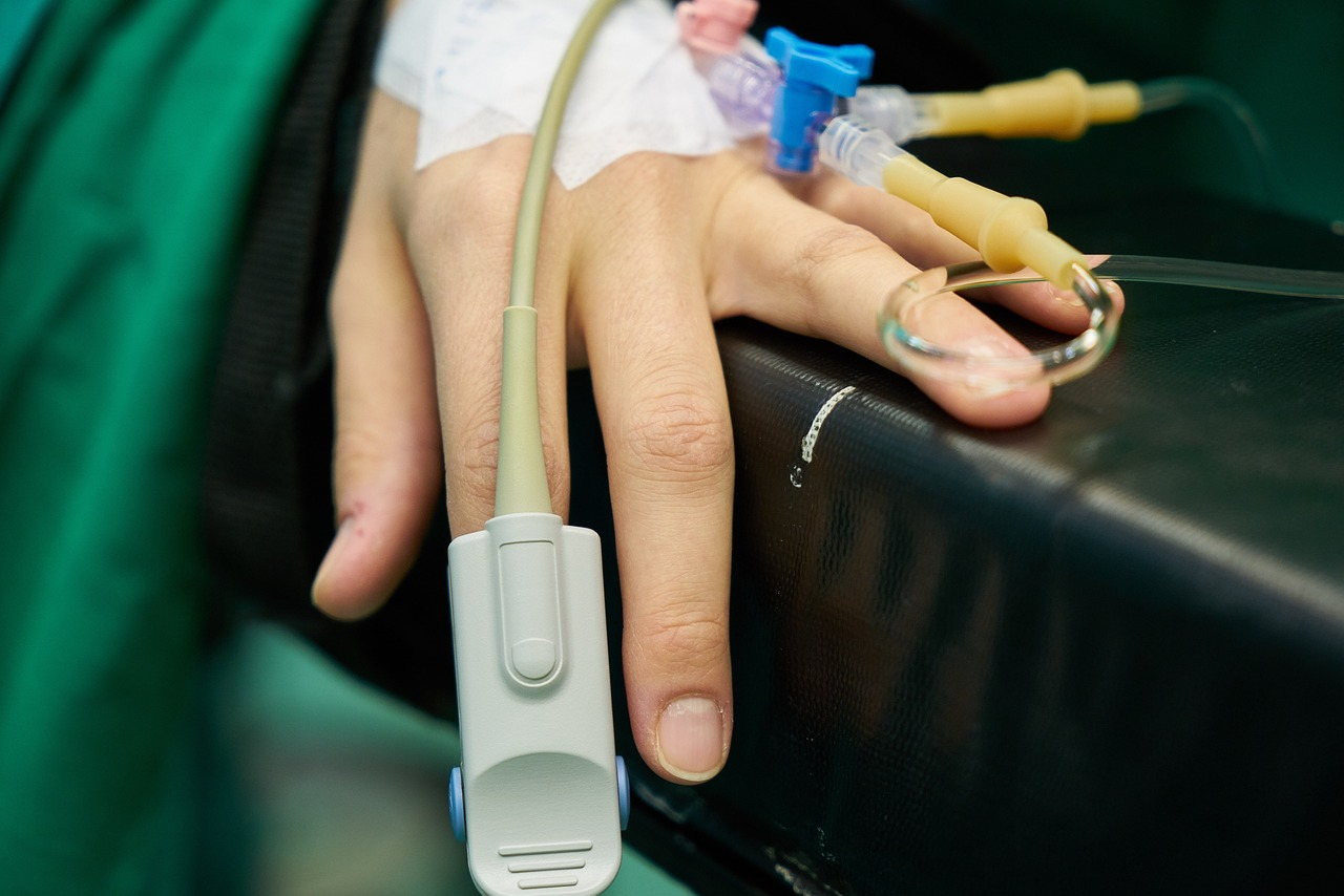 photo of a hand with a pulse oximeter on the index finger and an IV in the back of the hand