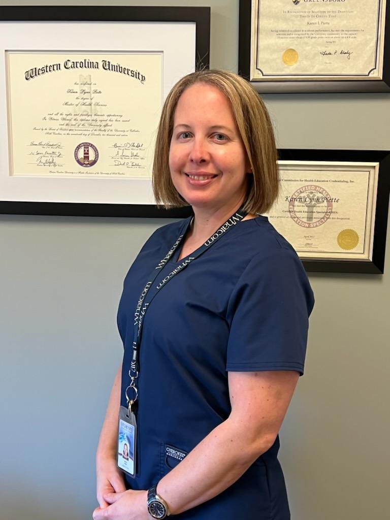 Karen Piette in medical scrubs smiling in front of a wall of her degrees and certifications