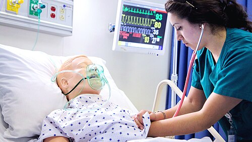 a nurse in a teal scrub top listens to a manikin’s chest with a stethoscope. The manikin represents a caucasian blonde male and has an oxygen mask on the face.