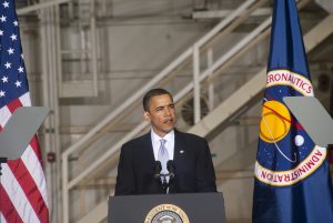 photo of 44th president of the United States Barack Obama speaking in front of a podium in front of an American flag, and a National Aeronautics and Space Administration flag.