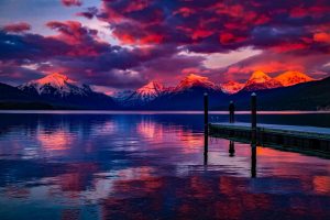Filtered pink and blue photo of sunset over a lake with a dock on the right side, mountains in the background, and fluffy clouds overhead.
