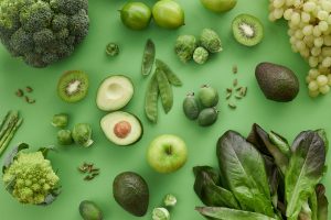Assortment of green fruits and vegetables on a green background to represent spring: broccoli, kiwi, avocado, limes, peas, an apple, brussel sprouts, asparagus, grapes, and lettuce.