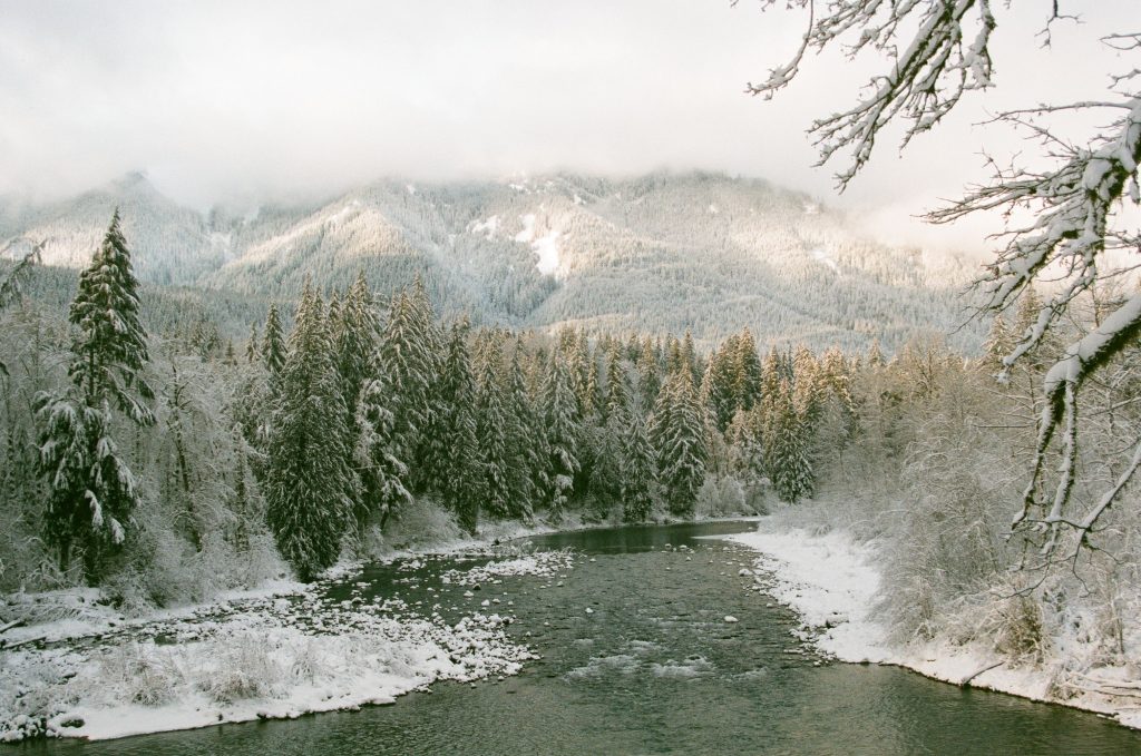 A snowy landscape overlooking an icy river lined with evergreens and bare trees. Mountains rise in the distance, disappearing into fog.