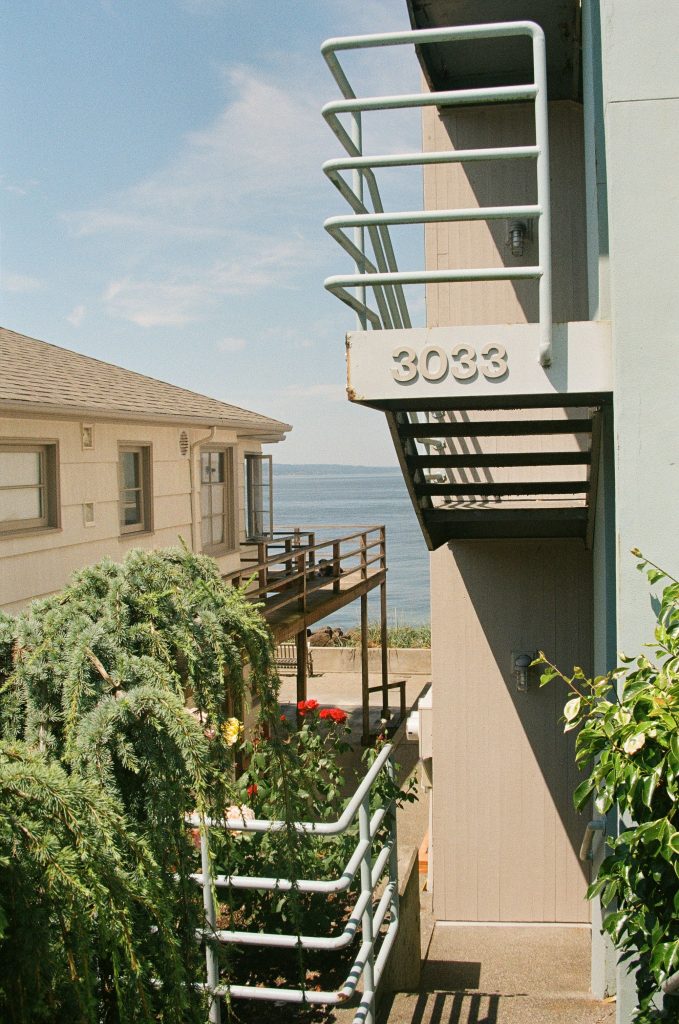 A two tiered apartment building with metal railing. '3033' is on the top railing while the bottom is lined with bright green bushes. There is a body of water and a clear blue sky in the background.