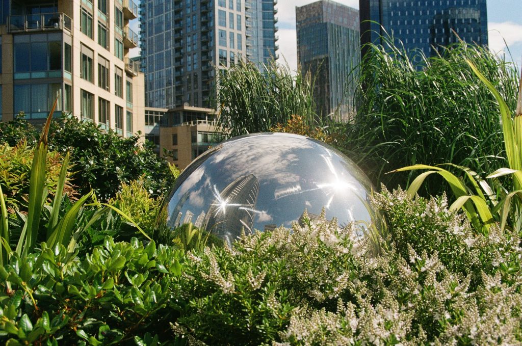 A silver metal sphere reflects tall buildings and sunlight. The sphere is surrounded by bright green plants. Buildings rise up behind the plants.