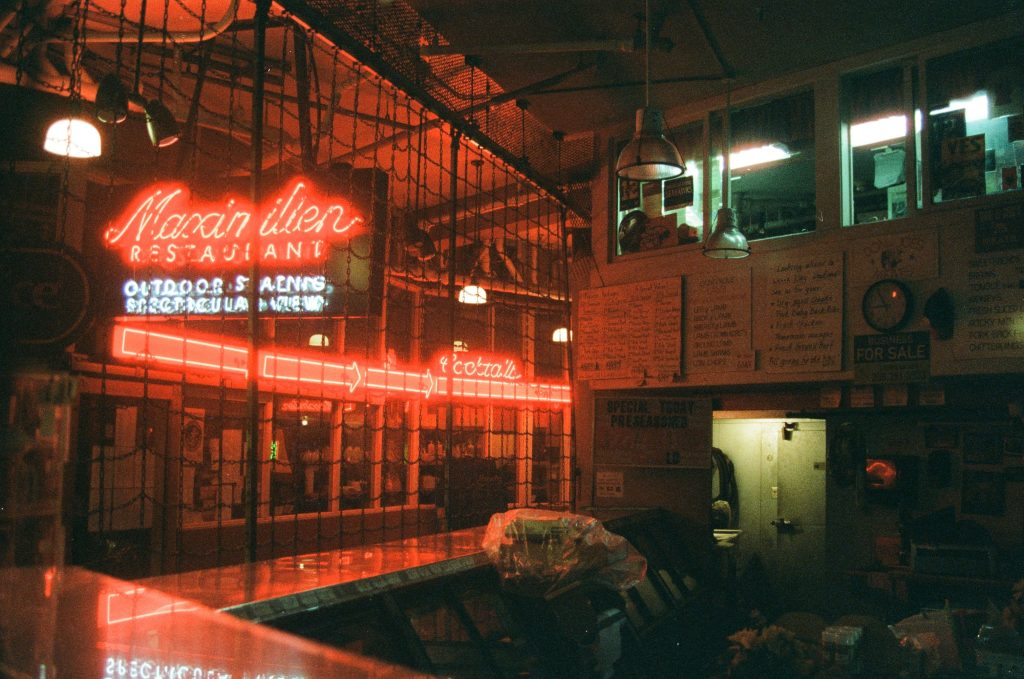 A dark, red lighted inside of an ice cream parlor, showing behind the counter while the parlor is closed. There are chains behind the counter, separating the parlor from the street. Across the street is a closed store, the name in neon red. Neon red arrows point to the right.