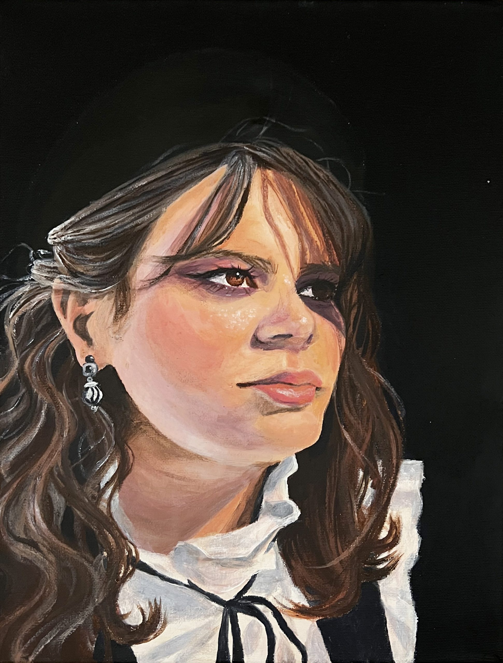 A portrait of a woman in a white blouse with a thing black ribbon tied in a bow around the collar. She is wearing silver earrings, winged eyeliner, and purple eyeshadow. Her brown wavy hair casts deep shadows across her face and she is against a matte black background.