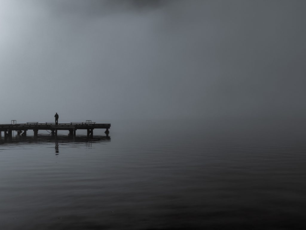 A vast body of water takes up the majority of the piece, fog hiding the horizon. There is a bridge in the midground reaching nearly to the middle of the page, with a silhouette of a figure standing on it. The piece is in grayscale and black.