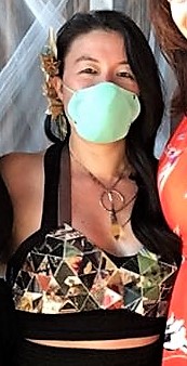 A woman wearing a mask and a pointed bra fashioned out of multicolor paper triangles over a black tank top. There are ribbon straps holding it in place tied behind her neck.