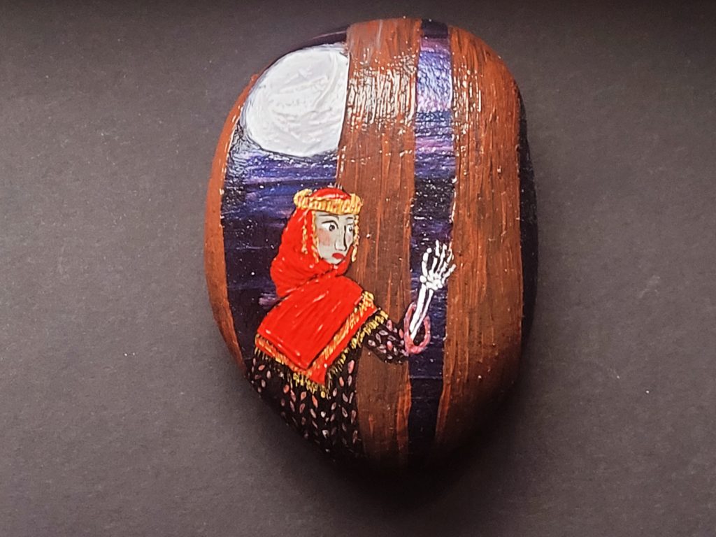A painted stone featuring a person in a red and gold shayla over a black dress with pink dots. They are lifting a skeletal hand and arm. Behind them are brown tree trunks in front of a dark purple sky. The moon can be seen between the trees.