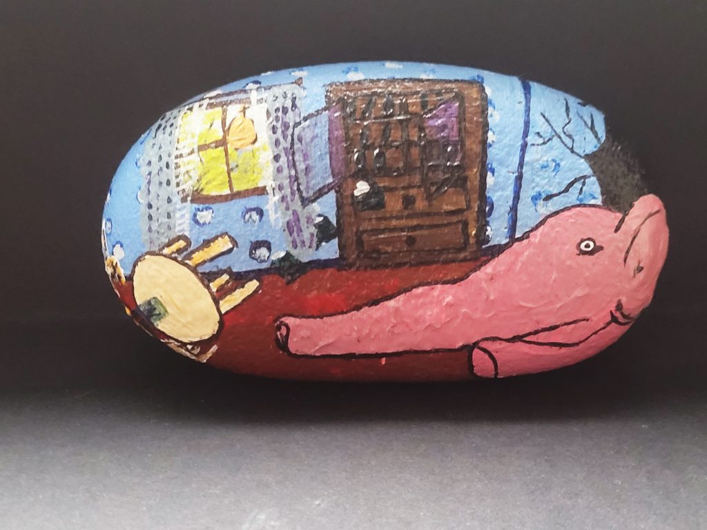 Painted stone with a large pink elephant's face and nose taking up most of the bottom. It is on a red carpet in front of a cabinet.  There is sunlight through a window and blue wallpaper.