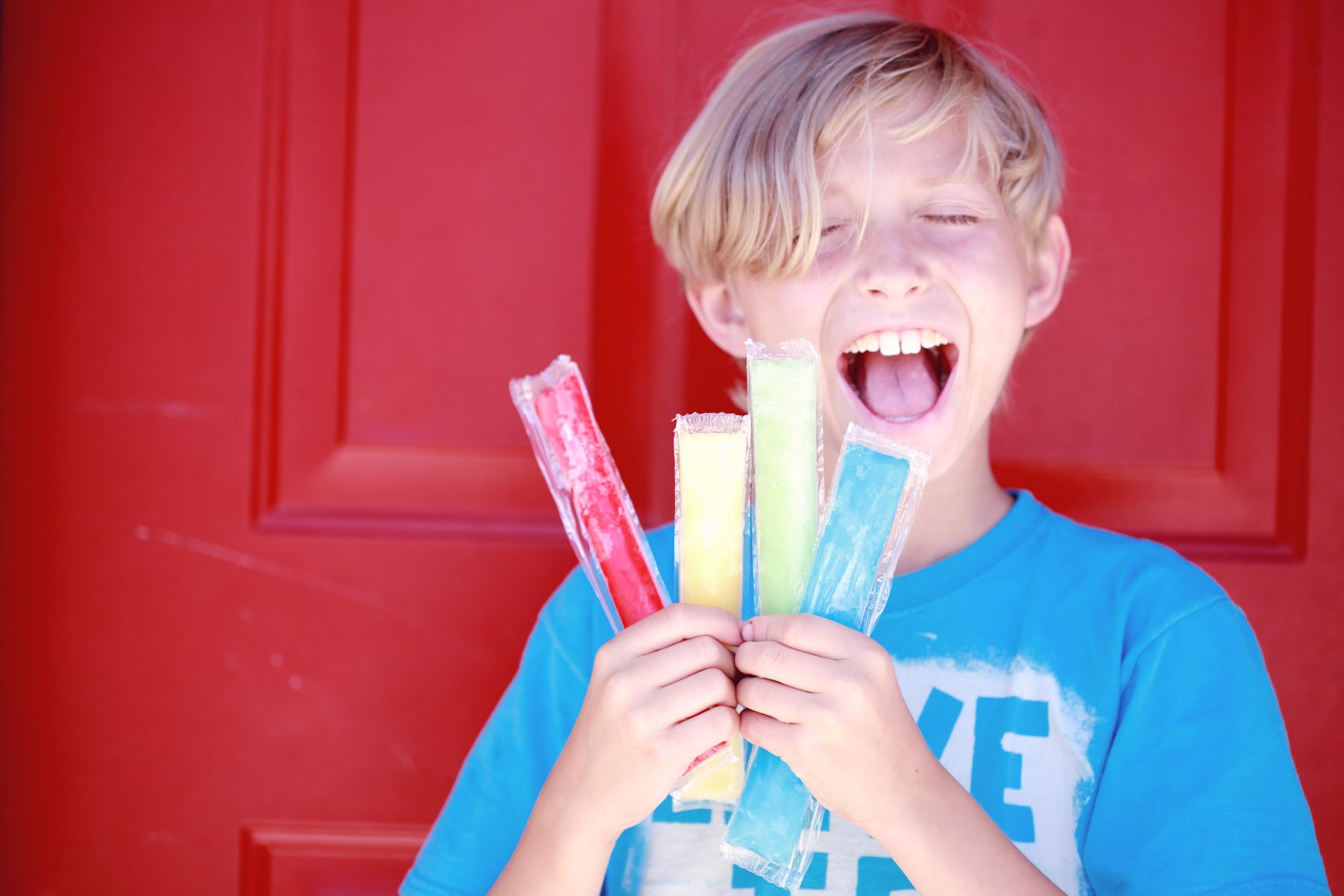 A young adolescent with fair skin and blond hair has a handful of frozen treats. He is smiling with his eyes closed and bringing a treat to his mouth.