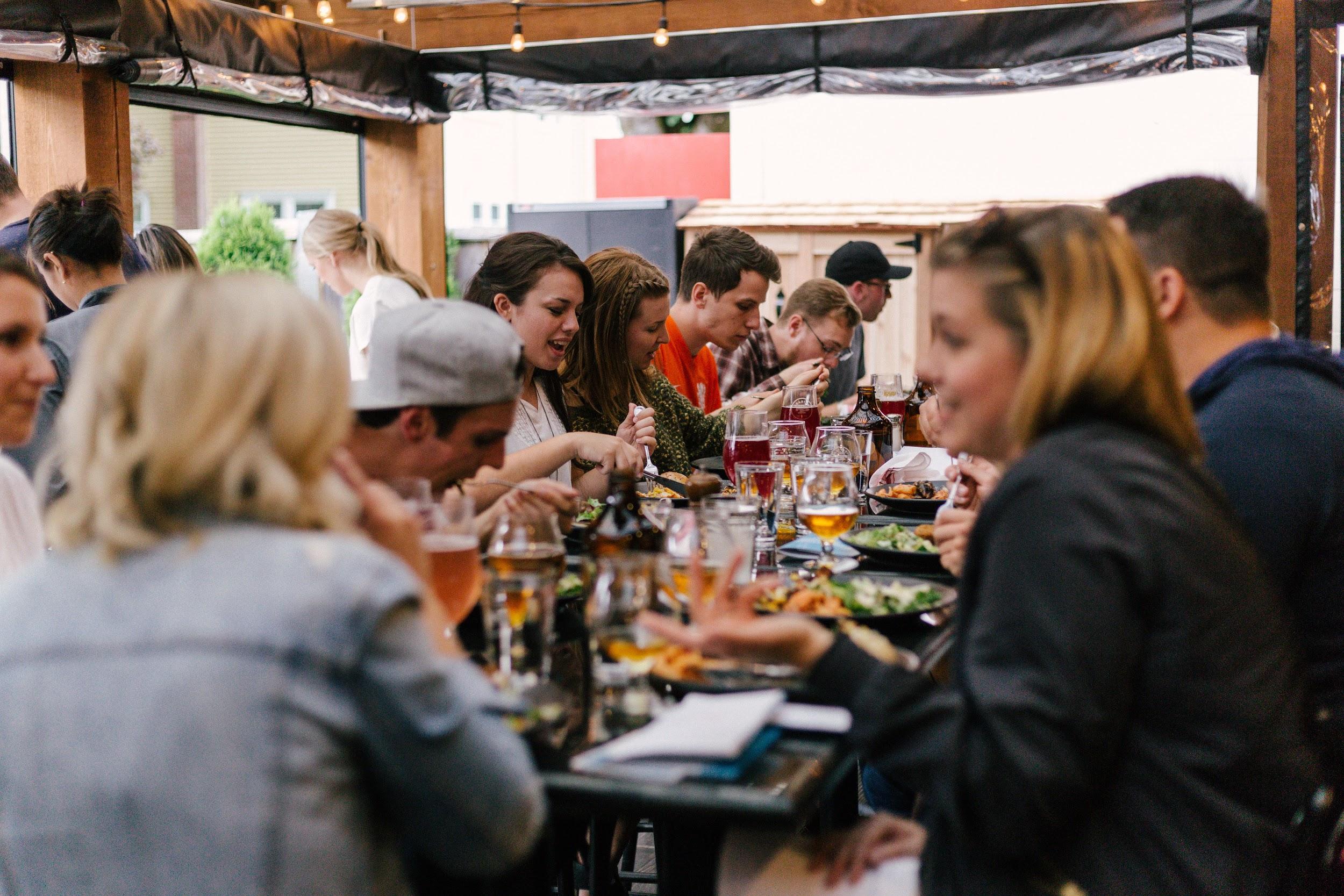 The image is a photo of a large group of people sharing a meal together in what appears to be an outdoor dining setting at a restaurant.