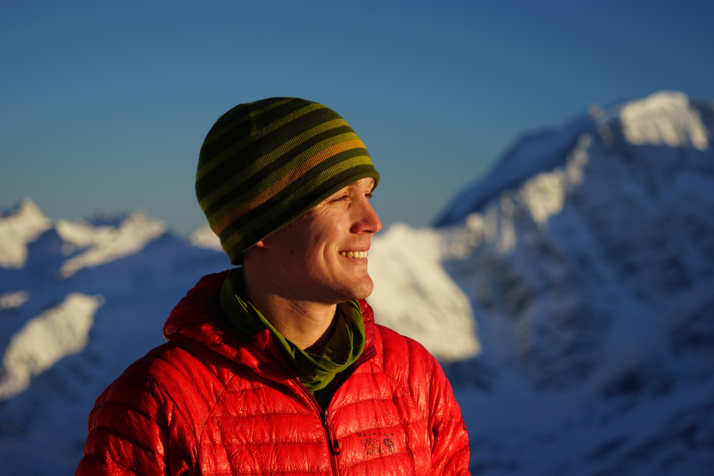 Colin O'Brady, a Caucasian man wearing a red hooded coat and a striped winter cap, looks off into the distance while sunlight shines on his face. In the background, snowy mountains are visible against a bright blue sky.