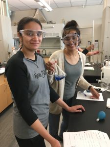 Image of two women in chemistry lab. They are standing at a lab table with supplies. Both are wearing goggles and smiling. One student is holding a beaker with a blue liquid.