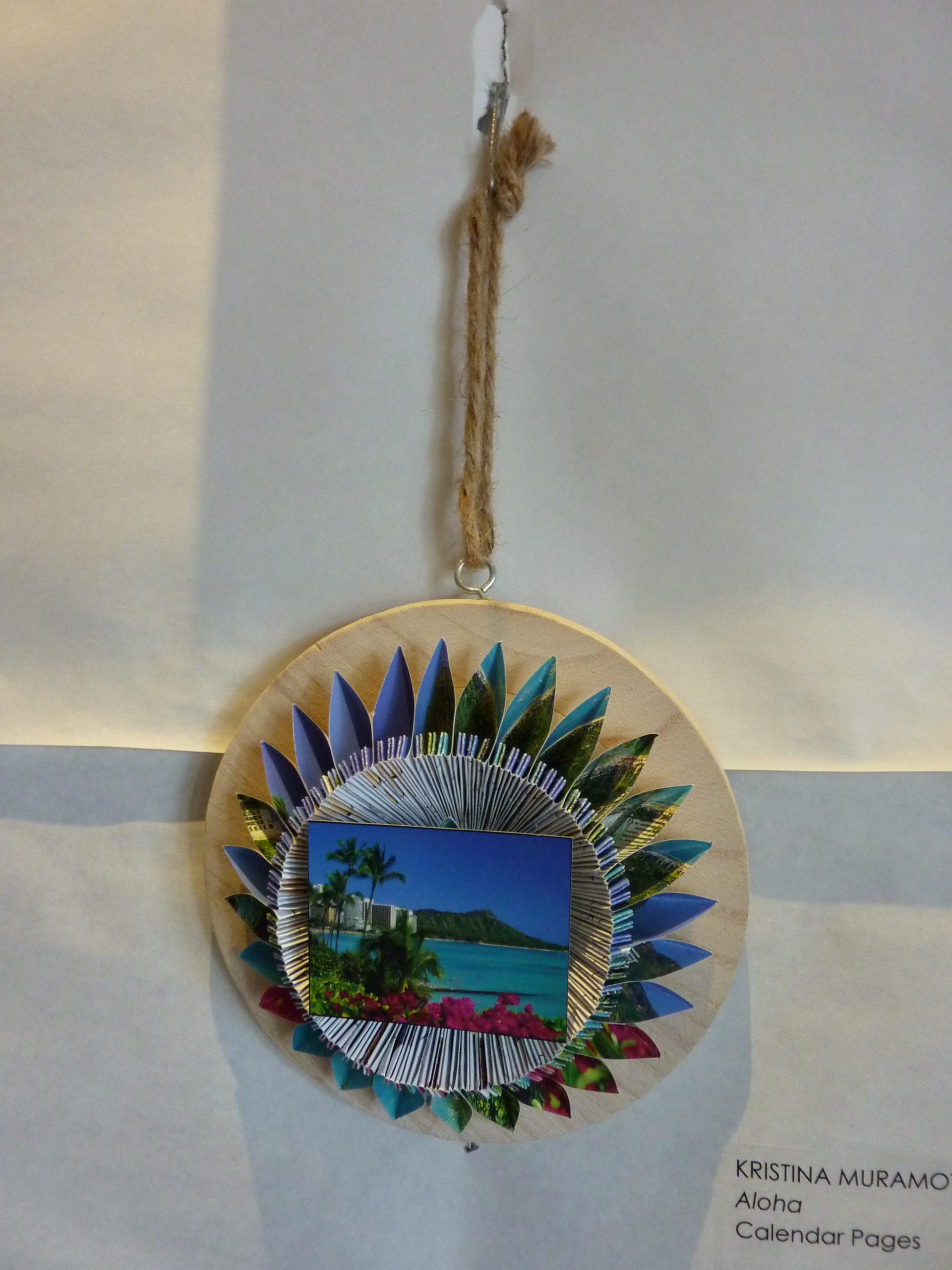 Wooden pendant with twine string with folded pieces of paper making a flower, and a postcard of a Hawaii beach in the middle