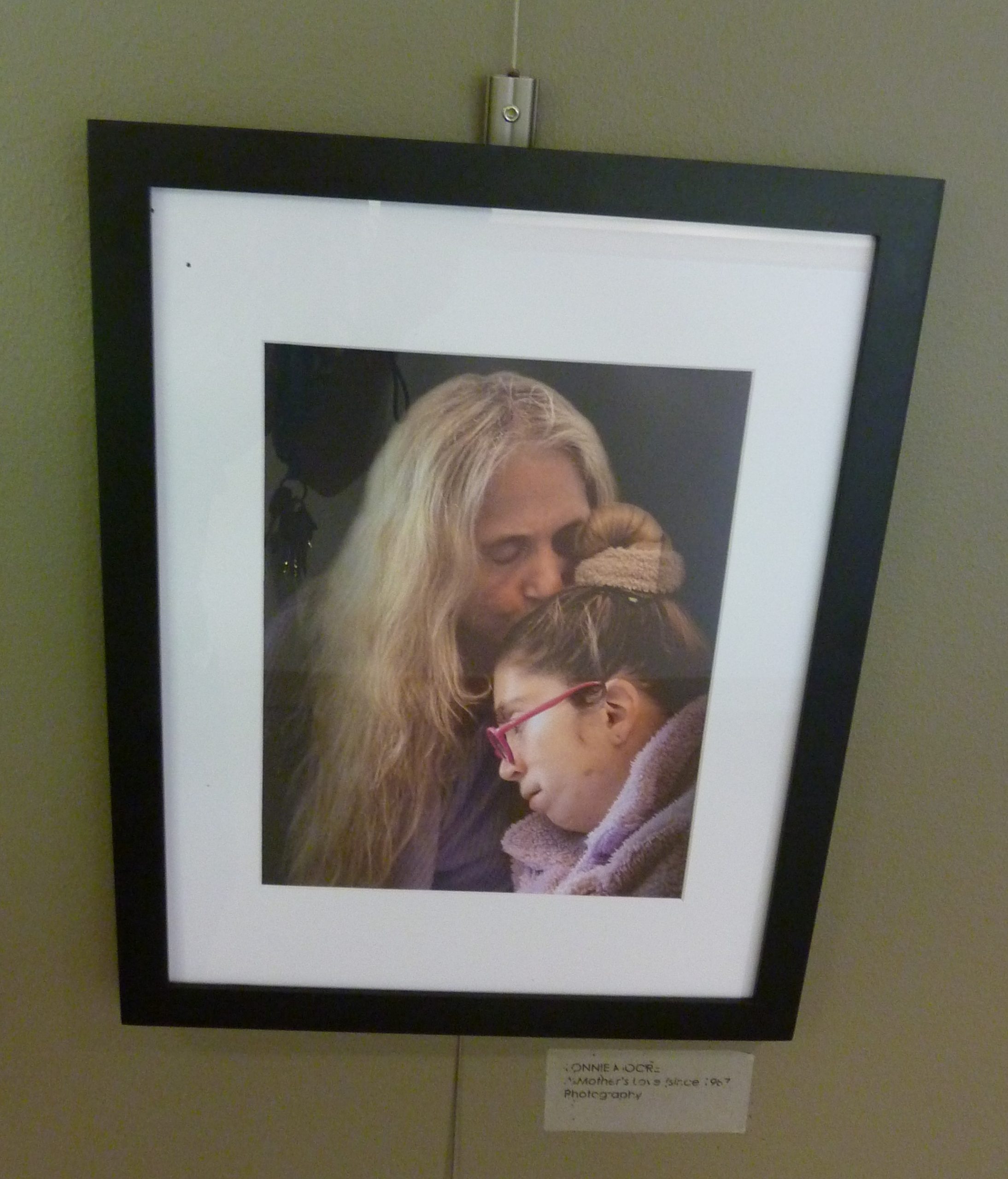 Color photograph of a person with long gray hair kissing the head of a person wearing pink glasses with a bun in their hair