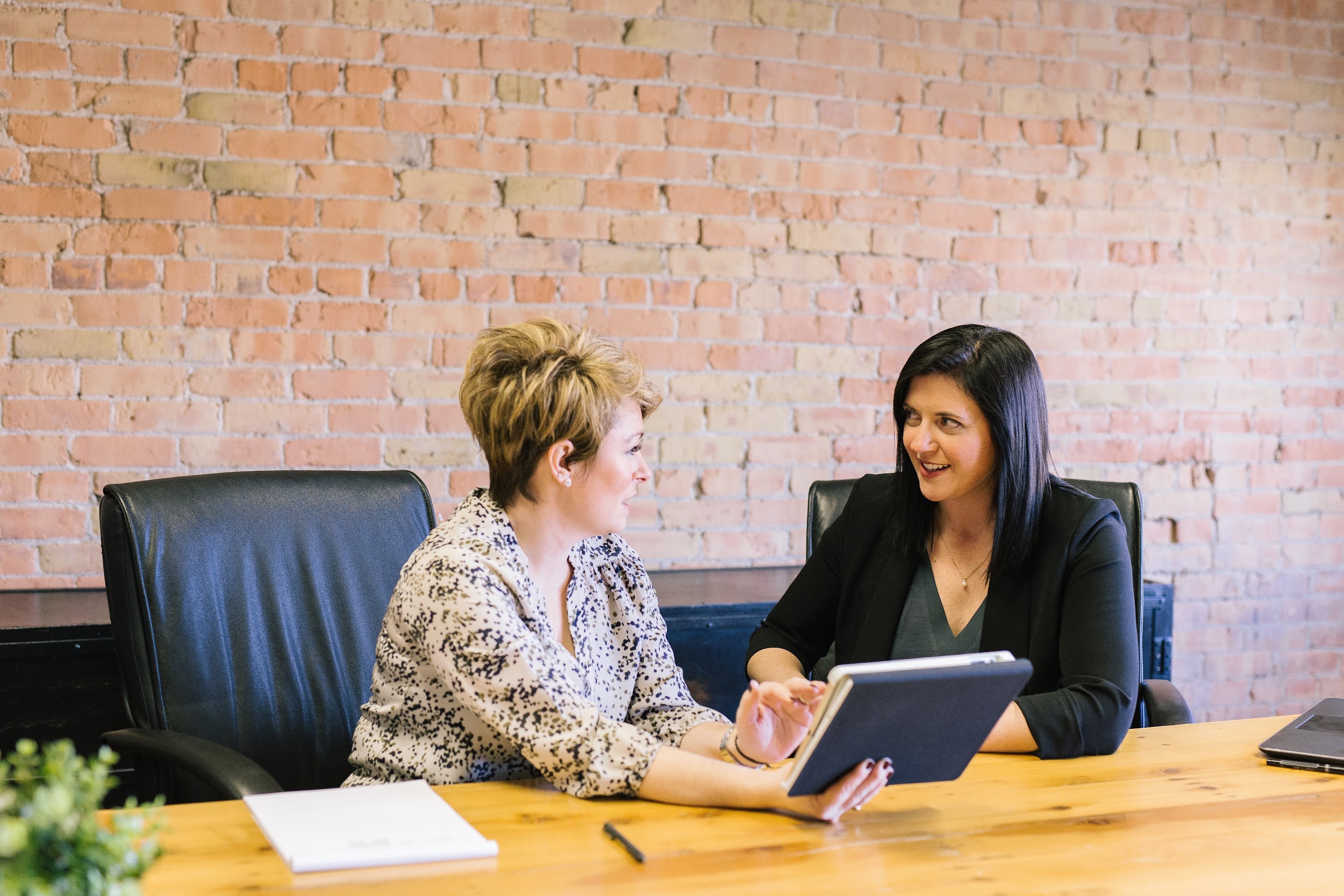 Two women sit at a conference table. One woman is showing some printed materials to the other woman and they are talking together.