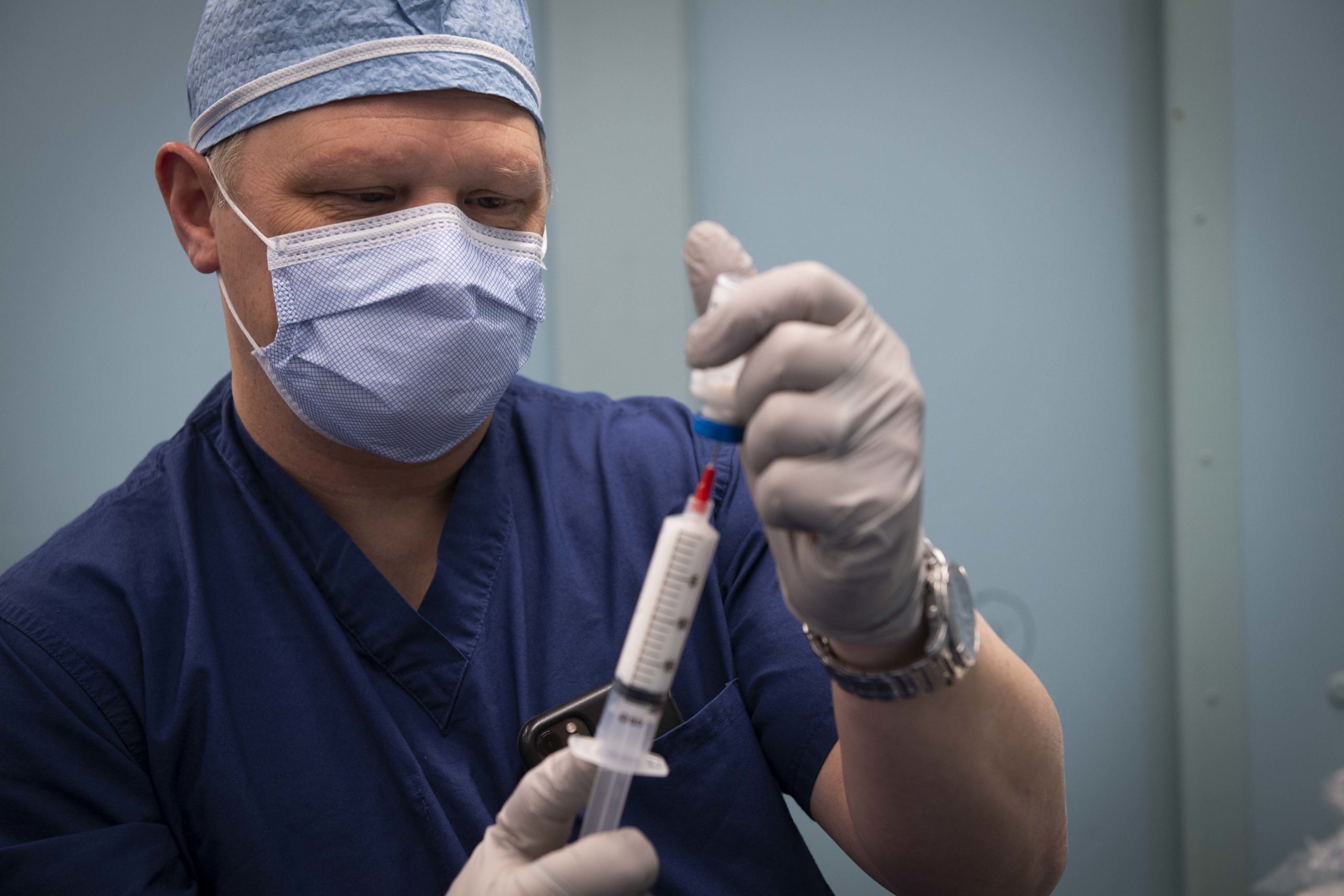 Photo showing man wearing surgical PPE, holding a syringe